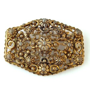 May include: A gold-toned metal brooch with a floral design. The brooch is made of intricate filigree work and features a variety of small, round, and flower-shaped details.