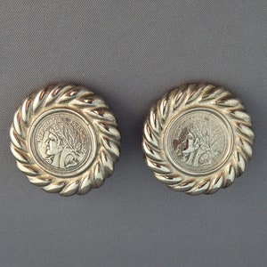 May include: Two silver-toned, round earrings. Each earring features a central coin design with a profile and the text "REPUBLIQUE FRANCAISE" surrounded by a twisted rope-like border. The earrings are set against a gray background.
