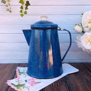 Puede incluir: Una cafetera de esmalte azul con un acabado moteado y una tapa. La cafetera está sentada sobre una servilleta blanca con un patrón floral. La cafetera está frente a un fondo de madera blanca.