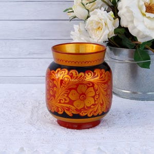 May include: A hand-painted, round vase with a black base and rim, adorned with a vibrant red and gold floral design. The vase has a gold interior and sits on a red base. White roses in a metal bucket are in the background.