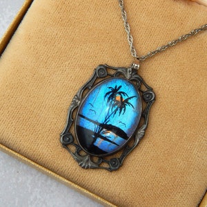 May include: Necklace with a silver-coloured pendant depicting a tropical scene. The oval pendant shows a blue sky, palm trees, and a sunset. The necklace is displayed in a gold-coloured box.