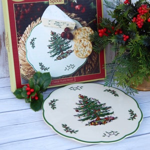 May include: A cream-colored Spode Christmas Tree cheese plate with a green scalloped edge. The plate features a detailed Christmas tree design with ornaments and gifts. The plate is displayed with a box, cheese, crackers, and holly.