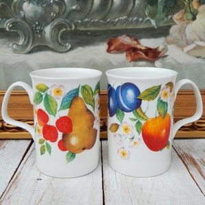 May include: Two white ceramic mugs with fruit designs. One mug features a pear, strawberries, and green leaves. The other mug displays plums and an orange fruit. Both mugs have a white handle and a floral border.
