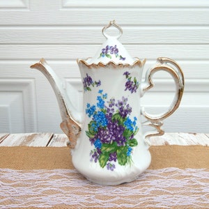 May include: White porcelain coffee pot with a lid, featuring a floral design in shades of blue and purple. The pot has a gold trim and handle, and a long spout. The pot is sitting on a burlap and lace surface.