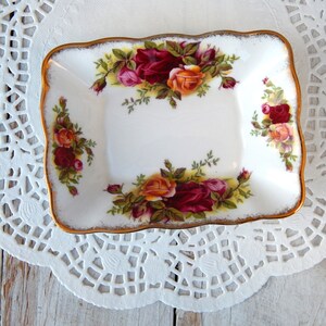 May include: A white, rectangular trinket dish with a scalloped edge and gold trim. The dish is decorated with a floral pattern of red, pink, and orange roses. The dish is placed on a white lace doily.