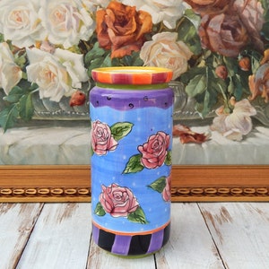 May include: A cylindrical ceramic vase with a blue background and pink roses painted on it. The vase has a purple, green, and yellow striped rim and a black and purple striped base.