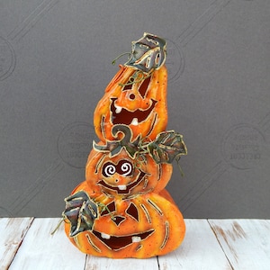 May include: A vertical stack of three orange metal jack-o'-lanterns with cut-out facial features, including wide grins and teeth. Each pumpkin is accented with dark green metal leaves and thin wire tendrils, featuring a distressed finish.