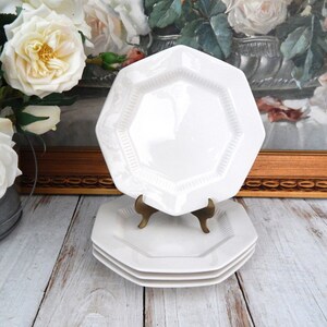 May include: A stack of three white octagonal plates with a decorative rim, one plate is displayed on a brass stand. The plates are on a light wood surface, with white roses in the background. The plates are a classic design.