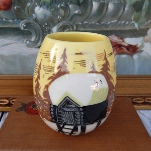 May include: A yellow ceramic vase with a hand-painted cabin scene. The vase features a cabin, trees, and a mountain landscape design. The vase is approximately 4 inches tall.