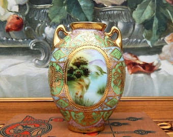 May include: An antique, hand-painted vase with a gold rim and handles. The vase features a landscape scene within an oval frame, surrounded by ornate gold detailing and pastel green and purple accents. The vase is approximately 6 inches tall.
