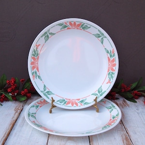 May include: Two white ceramic plates with a floral design. The plates have a border of coral-colored flowers and green leaves. One plate is on a gold-colored stand. The plates are on a white wooden surface.