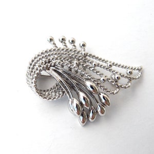 May include: A silver-toned brooch with a unique design. The brooch features a looped, twisted rope-like element, with a cluster of elongated, teardrop-shaped accents. Delicate beaded chains and small spheres add detail.