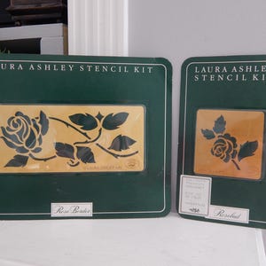 May include: Two Laura Ashley stencil kits. The kits are in dark green packaging with a gold-colored stencil design of a rose. The larger kit is labeled "Rose Border" and the smaller kit is labeled "Rosebud".