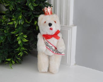 May include: A small, cream-colored teddy bear wearing a red and gold crown and a red ribbon sash. The sash reads "Berliner Bär" in black script. The bear has black eyes and a red mouth. The bear is standing on a white surface.