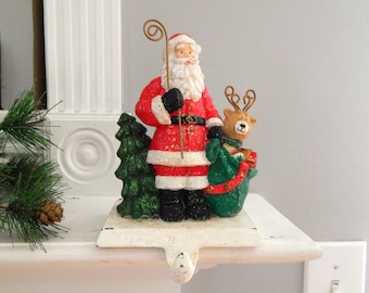 May include: A Christmas stocking holder featuring a Santa Claus figurine in a red suit with a white beard, holding a staff. A reindeer and green Christmas tree are also part of the design. The holder is white with a decorative hook.