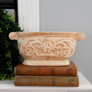 May include: A beige ceramic planter with ornate floral relief, featuring scrolled handles. The rectangular planter sits atop a stack of two antique books with worn, brown covers. Decorative home decor.
