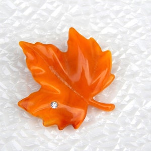 May include: Orange maple leaf-shaped decorative embellishment with a small, sparkling rhinestone. The leaf has a glossy finish and is set against a textured white background. Ideal for fall-themed crafts and decorations.