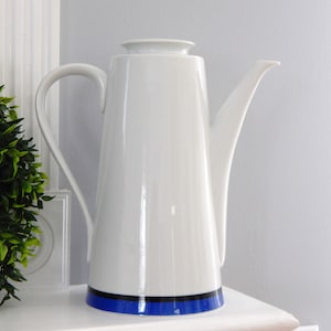 May include: A tall, white ceramic coffee pot with a black and blue band near the base. The pot has a long, curved spout and a handle. The design is minimalist and modern, suitable for serving coffee or tea. The pot is approximately 12 inches tall.