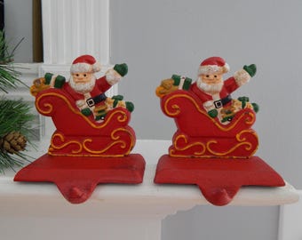 May include: Two red Christmas stocking holders, each featuring Santa Claus in a sleigh. Santa is dressed in a red suit with a white beard and hat. The sleigh is red with gold accents, and the holders are designed to hang stockings.