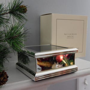 May include: A rectangular, silver-toned jewelry box with a glass top sits on a white shelf. The box has a mirrored finish and a decorative, ridged base. A box with text is behind the jewelry box. A pine branch with a pinecone is in the foreground.