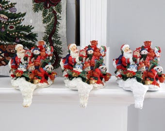 May include: Three Christmas stocking holders featuring Santa Claus and various festive characters. Each holder is a detailed sculpture with red, white, and green colors, designed to hang on a mantelpiece or shelf for holiday decor.