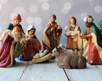 May include: A ceramic nativity scene featuring figures of the Holy Family, the Three Wise Men, and animals. The figures are painted in muted tones of burgundy, gold, and green. Baby Jesus lies in a manger, surrounded by a cow and sheep.