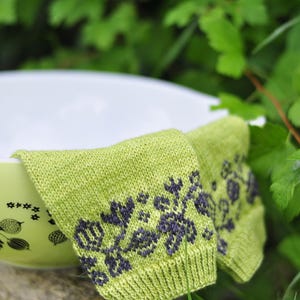 May include: A pair of green knitted socks with a floral pattern in a darker shade of green. The socks are folded and placed on top of a white bowl with a black and white floral pattern.