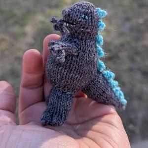 以下が含まれることがあります： グレーとブルーの色合いの、手編みの小さなゴジラのおもちゃ。編み物の恐竜は、灰色の体、青い背骨、小さな腕と脚を持っています。おもちゃは人の手で持たれています。