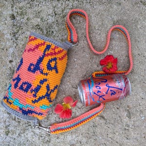 May include: A crocheted orange, yellow, and blue beverage holder with the text "La Croix" in blue. The holder has a long orange, yellow, and blue strap. A can of La Croix sparkling water is next to the holder.