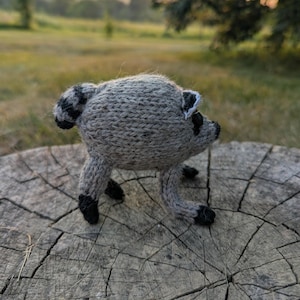 May include: A small, hand-knitted raccoon figurine crafted from heathered gray yarn. The creature features a rounded body, a striped tail, pointed ears with white accents, and black yarn detailing on the paws and facial features.