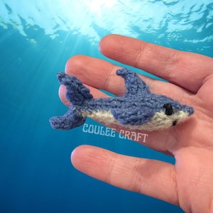 May include: A small, blue and white knitted dolphin toy held in a hand against a blue background. The dolphin has a white belly and black eyes.