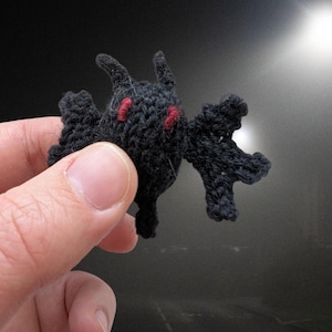 May include: A small, handmade black knitted creature with red eyes and small horns. The creature has outstretched wings and a stylized body. The background is a blurred image of a road and a bright light.