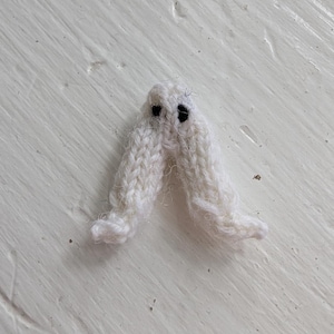 Op de afbeelding: Een kleine, handgemaakte, witte gebreide geest met twee zwarte ogen. De geest heeft een eenvoudig, minimalistisch ontwerp, perfect voor Halloween of griezelige decoraties. De geest is gemaakt van zacht, getextureerd garen.