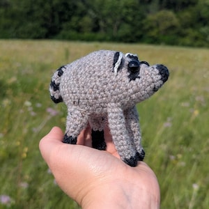 May include: A small, handmade crochet raccoon plush toy featuring a light grey body, black-tipped paws, a black nose, and a striped tail. The stuffed animal has round black plastic eyes and white accents around the ears, crafted with a textured yarn stitch.