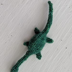 May include: A handmade, knitted green Loch Ness Monster toy. The toy features a long, curved neck, small flipper-like appendages, and a long tail. The toy is made of a textured yarn and is set against a white background.