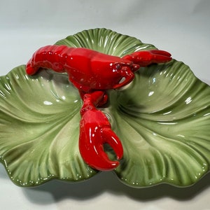 May include: A green ceramic serving dish shaped like a leaf with a red lobster figurine on top. The dish has three sections for serving different foods.