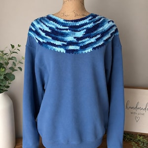 May include: A blue sweatshirt with a crocheted collar in shades of blue. The collar features a striped pattern with light and dark blue yarn. The sweatshirt is displayed on a mannequin.