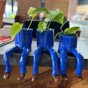 May include: Three quirky blue ceramic plant pots, designed to resemble denim trousers, each with brown shoes and holding green plants. The pots are displayed on a wooden surface, offering a unique and artistic design for home decor.