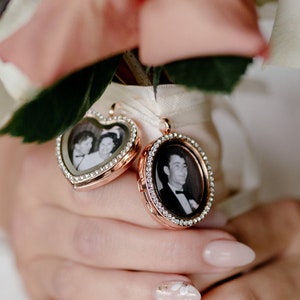 May include: A bride holds a bouquet of white and pink roses with two small gold locket charms attached. The lockets have photos of a couple inside.