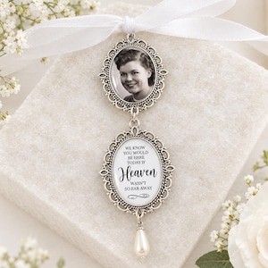 May include: A silver-toned metal memorial charm featuring two oval frames with ornate filigree edges. The top frame holds a black and white portrait of a person. The bottom frame contains the text: We know you would be here today if heaven wasn't so far away. A small white teardrop pearl hangs from the bottom.
