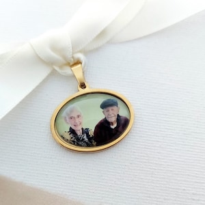May include: A gold oval-shaped pendant with a photo of an older couple. The pendant is attached to a white satin ribbon.