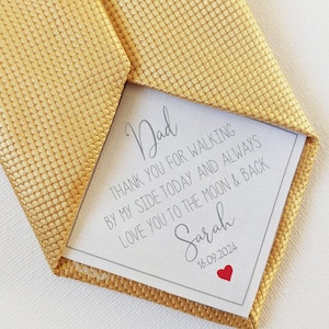 May include: A gold tie with a white card tucked inside. The card has a handwritten message that reads "Dad, Thank you for walking by my side today and always. Love you to the moon & back. Sarah 16.09.2024".
