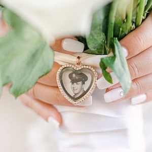 May include: A white bridal bouquet with a heart-shaped locket featuring a photo of a man in a military uniform. The locket is adorned with small, clear rhinestones.