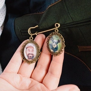 Peut inclure: Une broche dorée avec deux portraits de forme ovale. Les portraits sont encadrés de métal orné et représentent un homme et un chien. La broche est épinglée sur une veste vert foncé.