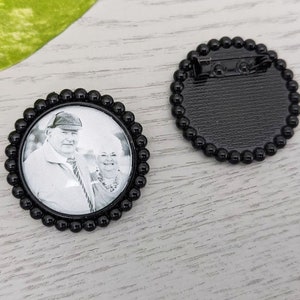 May include: Black and white photo of a couple in a round black frame with a beaded border. The back of the frame has a pin for attaching it to clothing.