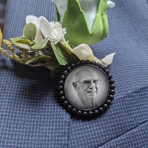 May include: A black and white photo of a man in a black frame with a white flower and green leaves attached to the frame. The frame is pinned to a blue suit jacket.