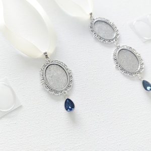 May include: Two silver pendant charms with ornate frames and clear glass inserts, each with a blue teardrop crystal hanging below. The charms are attached to white satin ribbon.
