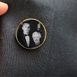 May include: A gold-toned pin with a black and white photo of a couple. The man is wearing a tuxedo and the woman is wearing a dress.
