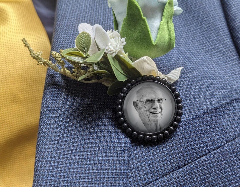 May include: A black and white photo of a man in a black frame with a white flower and green leaves attached to the frame. The frame is pinned to a blue suit jacket.