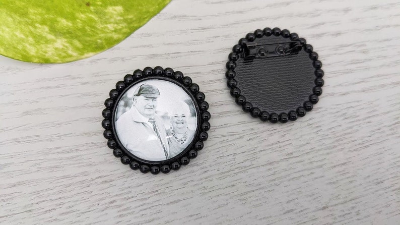 May include: Black and white photo of a couple in a round black frame with a beaded border. The back of the frame has a pin for attaching it to clothing.